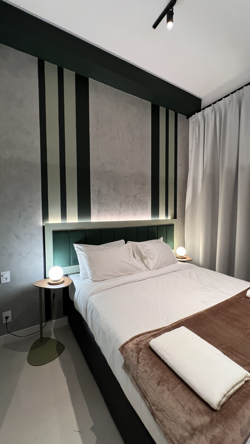 Área de dormir de studio com cama branca, cabeceira verde estofada, parede geométrica e luminárias esféricas laterais.